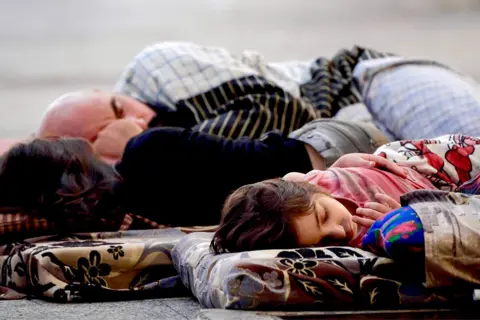 Darren Conway / BBC Father and  two children sleep outside on thin bedding.