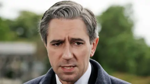 PA Media Simon Harris is speaking to the media. He has salt and pepper coloured hair and brown eyes. He's wearing a grey coat and a black suit with a white shirt and a red and white tie. He's standing outside. The background is blurred but trees and foliage can be seen.