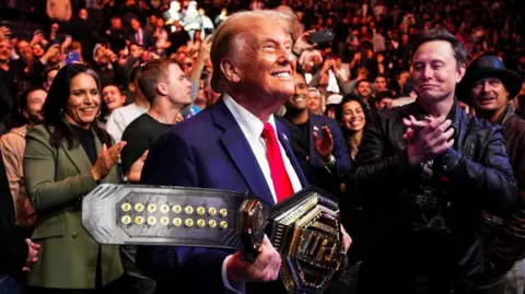 Getty Images Donald Trump grins while holding a UFC belt handed to him by UFC fighter Jon Jones. He is surrounded by a crowd including Elon Musk and Tulsi Gabbard. 