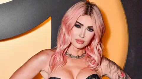 Getty Images Megan Fox staring at a camera. Wearing a gold necklace, with pink hair and tattoos on her left arm. She's stood in front of an orange and black background. 