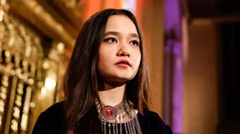 Getty Images Nila Ibrahimi stands in front of a gold and purple background, wearing a dark top and a necklace with red colouring