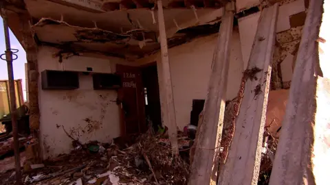 The Matías family house has been largely reduced to rubble, though some walls are still standing. There is debris on the floor and there is a door with the boys' names on that is still standing.