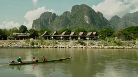 BBC Two tourists sit in a green and red canoe along a river, against a backdrop of jagged wooded peaks, tourist huts and a cloudy blue sky. 