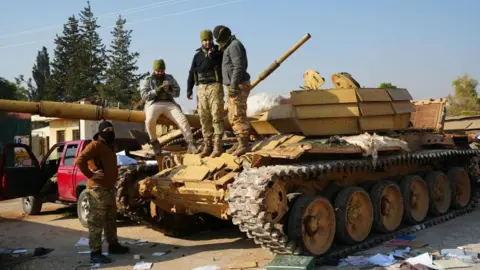 Getty Images Three men stand on top of a large yellow army tank, while one stands on the ground nearby. 