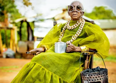 Luxury Media Zambia A seated Margret Chola wearing a chunky gold chain over a green taffeta like dress with puffy sleeves and a big frill for the hem holds a tin mug in her hand. Hanging off the arm of her chair is a "mbaula" filled with charcoal. She is wearing ornate framed sunglasses, the left eye frame in the shape of a D and the right frame in the shape of a C.