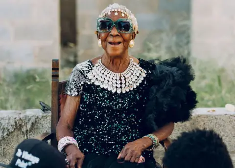 Kooma Jnr A seated Margret Chola laughs at the camera. She is wearing an Indian-style  head band, big bejewelled green-framed sunglasses, a diamond-like waterfall necklace and a black feather boa over a top covered in sequins