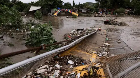 Reuters Twisted metal and debris can be seen partially submerged in the flooded waters of the Bermejo river. On the other side of the river's bank, people are standing and watching. 