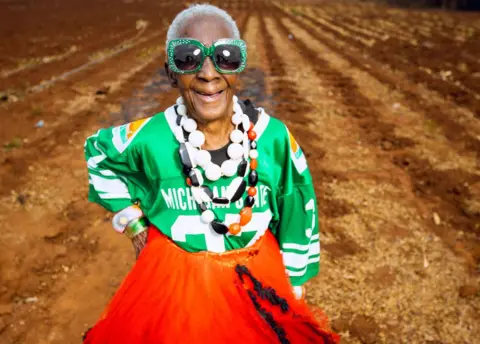 Luxury Media Zambia Margret Chola smiling in a field with her hand on her hip, wearing big green bejewelled sunglasses, a green sports jersey, a red dress and big plastic white, red and black beads around her neck.