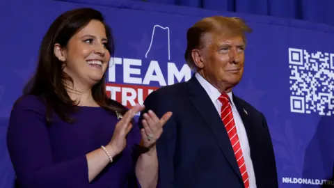 Getty Images Elise Stefanik wears a blue dress and claps her hands. She is standing on a stage next to Donald Trump, who wears a dark suit and red tie.