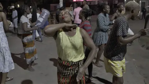 A woman with a whistle holding up her hand across her chin - around her are other protesters holdings pots, bottles or whistles on a street at night in Maputo, Mozambique