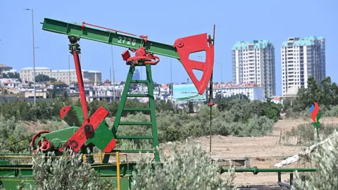 Getty Images A red and green oil pump at an oil well in the capital city Baku, with apartment blocks, a screen and a park in the background 
