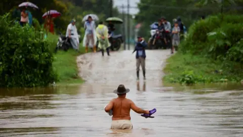 Reuters A shirtless man wearing a brown hat and holding his flip flops in one hand wades across waist-deep water while people watch from the other side of an inundated road in El Progreso, Honduras.