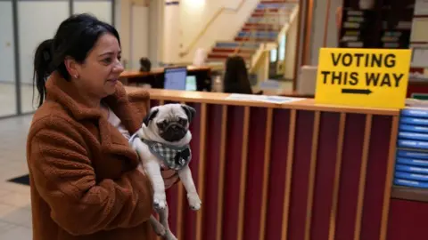 PA Media A woman holds a pug as she passes a yellow 'Voting this way' sign