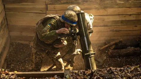 Getty Images Two Ukrainian soldiers lean over a mortar in a wooden ditch preparing to fire at Russian positions in the Sumy region on 30 September 2024