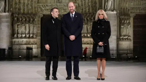 Reuters President Macron, wearing a black overcoat and scarf with a white shirt, says something to Prince William, who is also wearing a black overcoat and a white shirt with a blue tie. Brigitte Macron, wearing a black coat, black scarf and beaded black bag looks on beside them as they stand outside the Notre Dame 