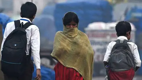 Getty Images A woman covers her face as she walks past school boys on a cold smoggy morning in the old quarters of New Delhi in November 2024