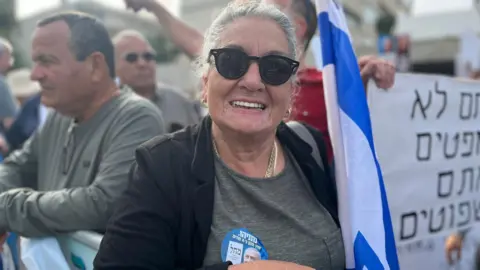 Eliza Ziv, a woman with grey hair and round black sunglasses, smiles at the camera. She is holding a folded Israeli flag and wears a sticker with Netanyahu's face on it.