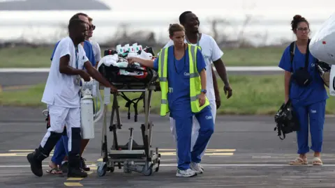 Reuters Paramedics in scrubs walk along a runway while pulling a stretcher carrying an injured person to an aircraft