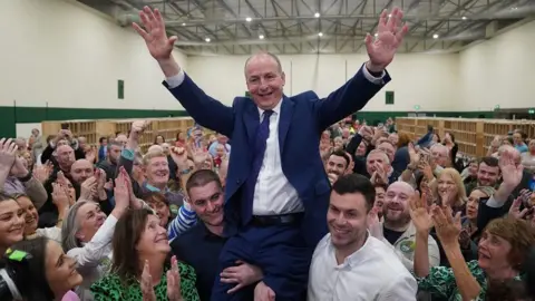 PA Media Micheál Martin who has balding light brown hair and is wearing a navy suit and white tie  is lifted up on the shoulders of two men while supporters clap and cheer