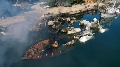 Getty Images A sunken large boat close to the dockside in the Syrian port town of Latakia. There is smoke rising and partially submerged metallic structures. 