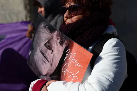 Reuters A woman wearing a white coat and sunglasses holds a sign with Gisèle Pelicot's face with the words "merci Gisèle".