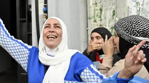 EPA A smiling woman in a blue and white dress looks up to the sky. Behind her two other women cover their smiles with their hands.