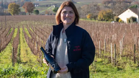 Coralie de Bouard stands in a vineyard holding a bottle of her wine