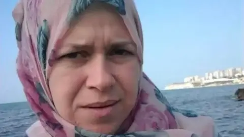 Sanaa Azzam Ghinwa Muhammad Azzam looking into the camera, she is wearing a pink and blue floral headscarf. The sea and coastal buildings are in the background.