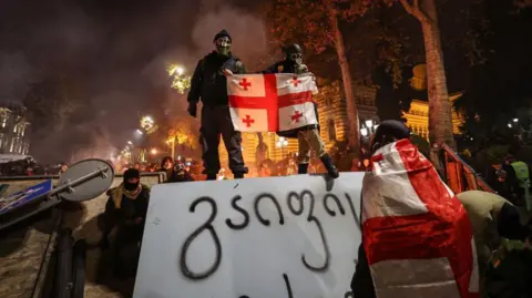 GIORGI ARJEVANIDZE/AFP A protester wearing a Georgian flag stands on a barricade during a clash with police on December 1, 2024