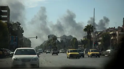 Getty Images Smoke rising on the horizon of a street in Damascus, Syria. Cars are driving down the road. It is day time with a blue sky.