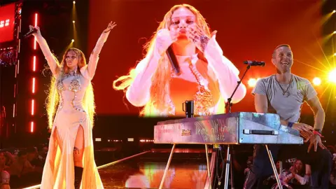 Getty Images Elyanna and Chris Martin of Coldplay perform onstage at the 2024 iHeartRadio Music Festival in Las Vegas. Elyanna is wearing a long light coloured outfit and has her arms outstretched towards the sky. Her image is on the big screen behind her, while Chris Martin is sitting at a silver coloured keyboard, wearing a grey tshirt, smiling and looking out to the crowd.