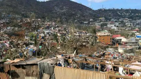 Getty Images Damage in Mayotte. A town is seen in ruins, with a hill scape rising in the background. Trees appear blown over, walls are collapsed, though some buildings remain standing.