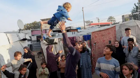 Reuters A man throws a child into the air at a camp in central Gaza for displaced Palestinians, following the beginning of the ceasefire on Sunday