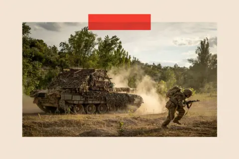  A -80 tank covered in camouflage fires while training, and a soldier with a gun runs from the vehicle. 