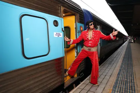 Don Arnold/WireImage An Elvis impersonator wearing in a red jumpsuit points to the sky as he steps on to a train.