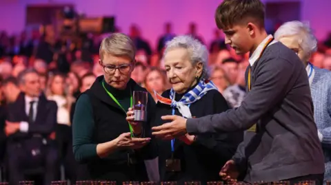 Beata Zawrzel/NurPhoto An elderly woman who survived the Birkenau death camp is helped by a younger woman and a young man to place a candle