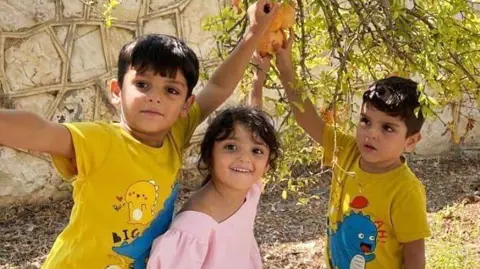 Fares family Hawraa, and her cousins Hassan and Hussein, photographed playing together. Hawraa is wearing a pink dress with puff sleeves and a square neck line. Her cousins are both in yellow cartoon dinosaur t-shirts.