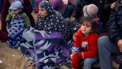 Reuters Palestinians wait to be allowed to return to their homes in northern Gaza after they were displaced to the south at Israel's order during the war. A woman in a headscarf sits with a blanket on her knees, with a child wearing a red jumper next to her.