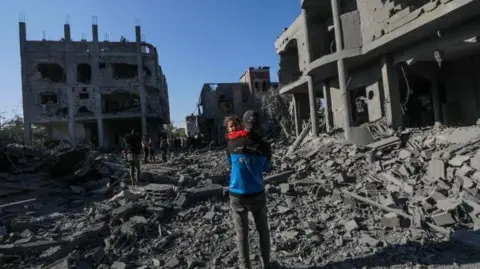 EPA A Palestinian father carries his daughter next to a destroyed house in Al Maghazi refugee camp, central Gaza Strip on 03 January 2025.