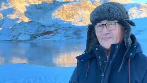 Kaaleeraq Ringsted wearing a black jacket and hat smiling in front of some fish with a fjord in the background