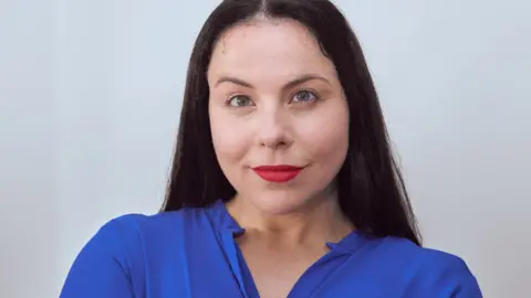 Amie Liebowitz A woman with long dark hair is smiling at the camera. She is stood in front of a white background and is wearing red lipstick and a blue blouse.