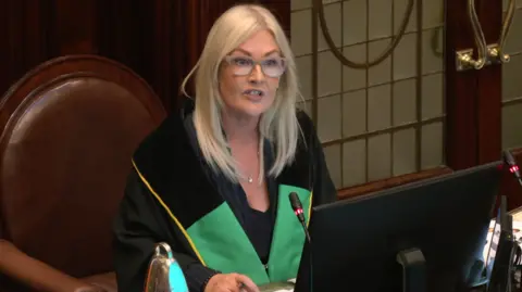 Verona Murphy, who has long blond hair and is wearing light-framed glasses, looks towards members of the Dail while sitting at her desk. She is wearing a green and black robe and a microphone is in front of her.