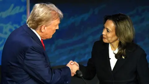 AFP Donald Trump and Kamala Harris shake hands while smiling courteously at each other, both wearing suits, against a blue backdrop on stage in Philadelphia in September