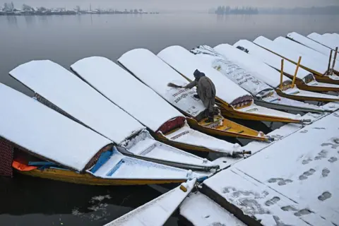 TAUSEEF MUSTAFA/AFP A row of boats covered in snow on a misty lake in India. A man wearing grey uses a brush to remove the snow