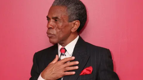 Lia Chang André De Shields, with streaks of grey hair, holds his hand against his charcoal pin-stripped jacket and tie. A red handkerchief pokes out of a pocket. He is pictured against a red wall.