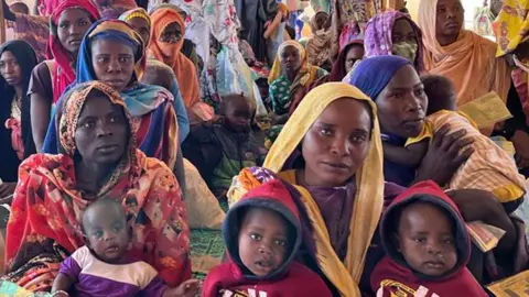 Women in colourful headscarves sitting on mats, some clutching children on their laps at a makeshift reception area at the Adré border post in Chad