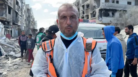 Haitham al-Homs, a man wearing an orange high-vis item and in protective forensic gear, stands in front of an ambulance in Rafah