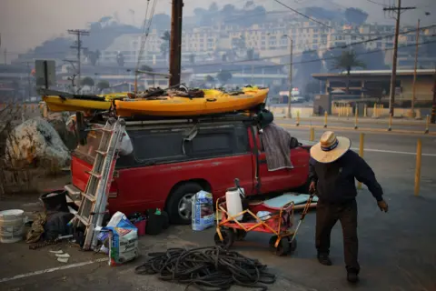 Daniel Cole/Reuters A man in a wide-brimmed hat fetches water while standing next to a red vehicle with a kayak on top of it, along the Pacific Coast Highway on Wednesday.