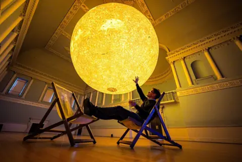 Ben Birchall/PA Luke Jerram sits in a chair and raises his hand towards his latest astronomical sculpture, Helios
