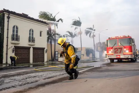 Allison Dinner/EPA A Los Angeles County firefighter drags a water hose in front of a fire engine that's shooting water at a building, as they take up positions to battle the Palisades wildfire in Malibu on Wednesday.